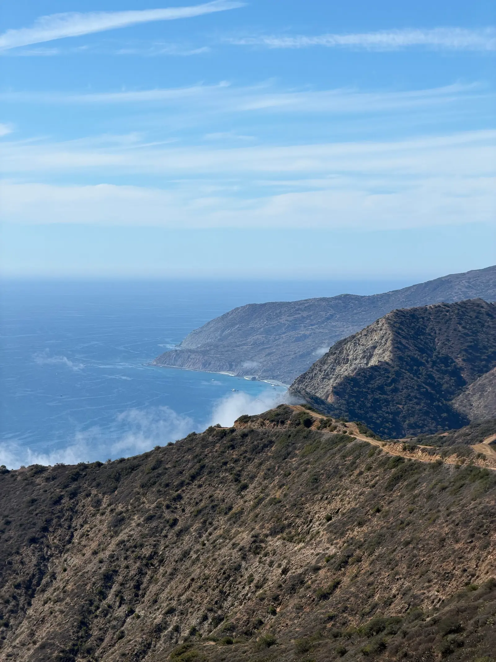 A view of the coastline showing rugged mountains descending into the sea with wisps of fog.