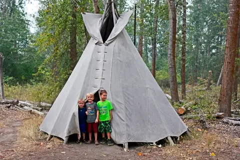 Three children stand in front of a teepee located in a wooded area.