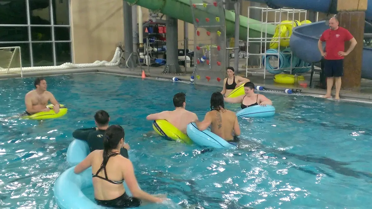 A group of adults in a swimming pool with inner tubes are being observed by a man standing on the pool deck.