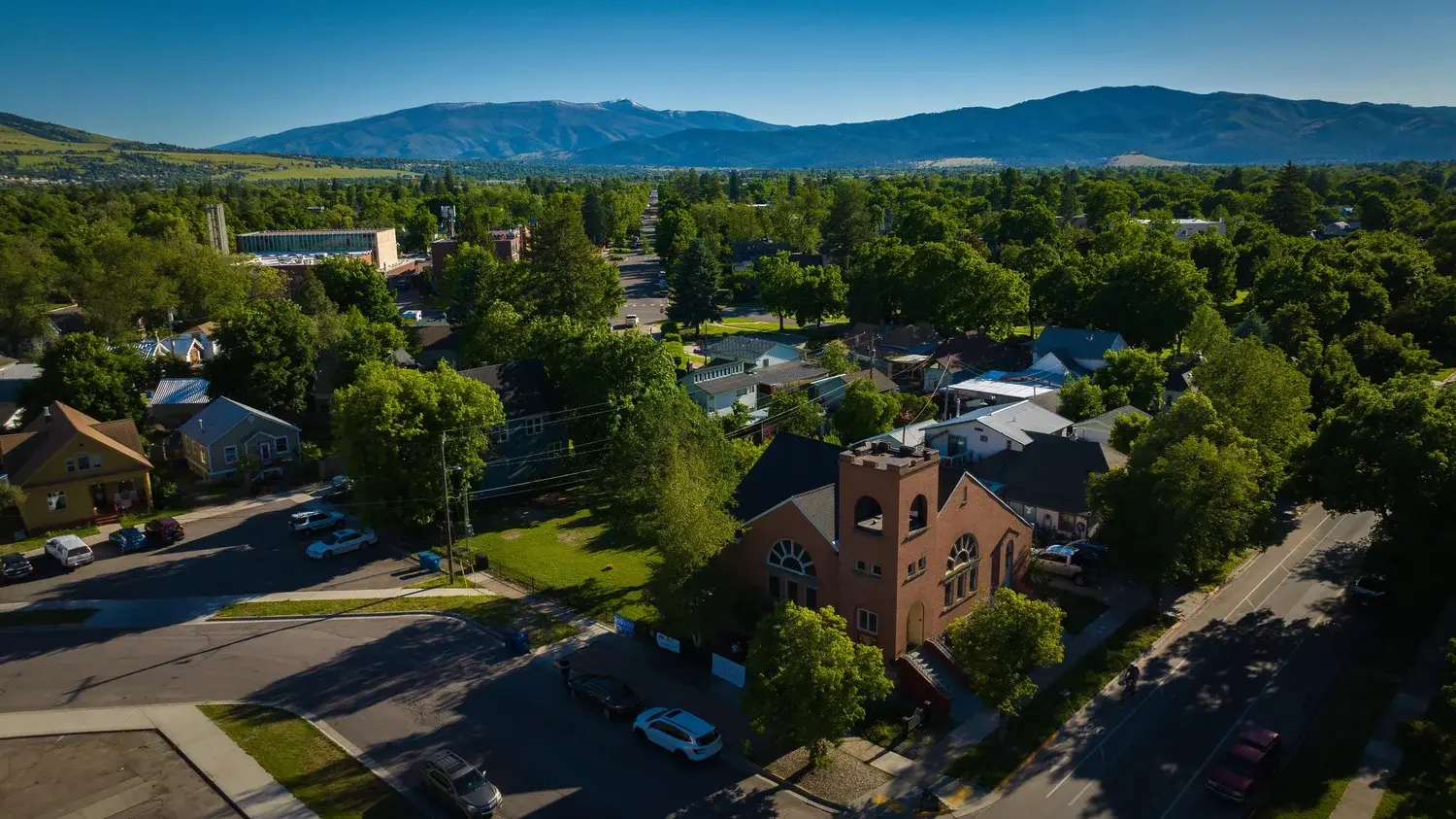An aerial view shows a brick church situated in a neighborhood surrounded by trees with mountains in the background.