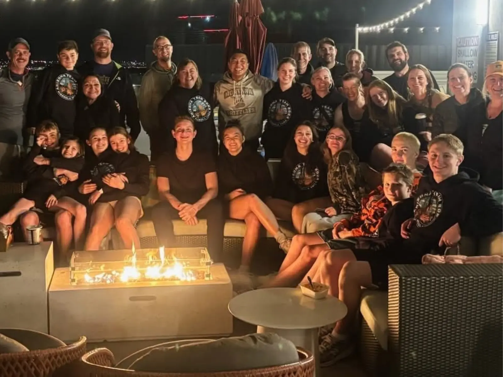 A large group of people are gathered together sitting around an outdoor fire pit at night.