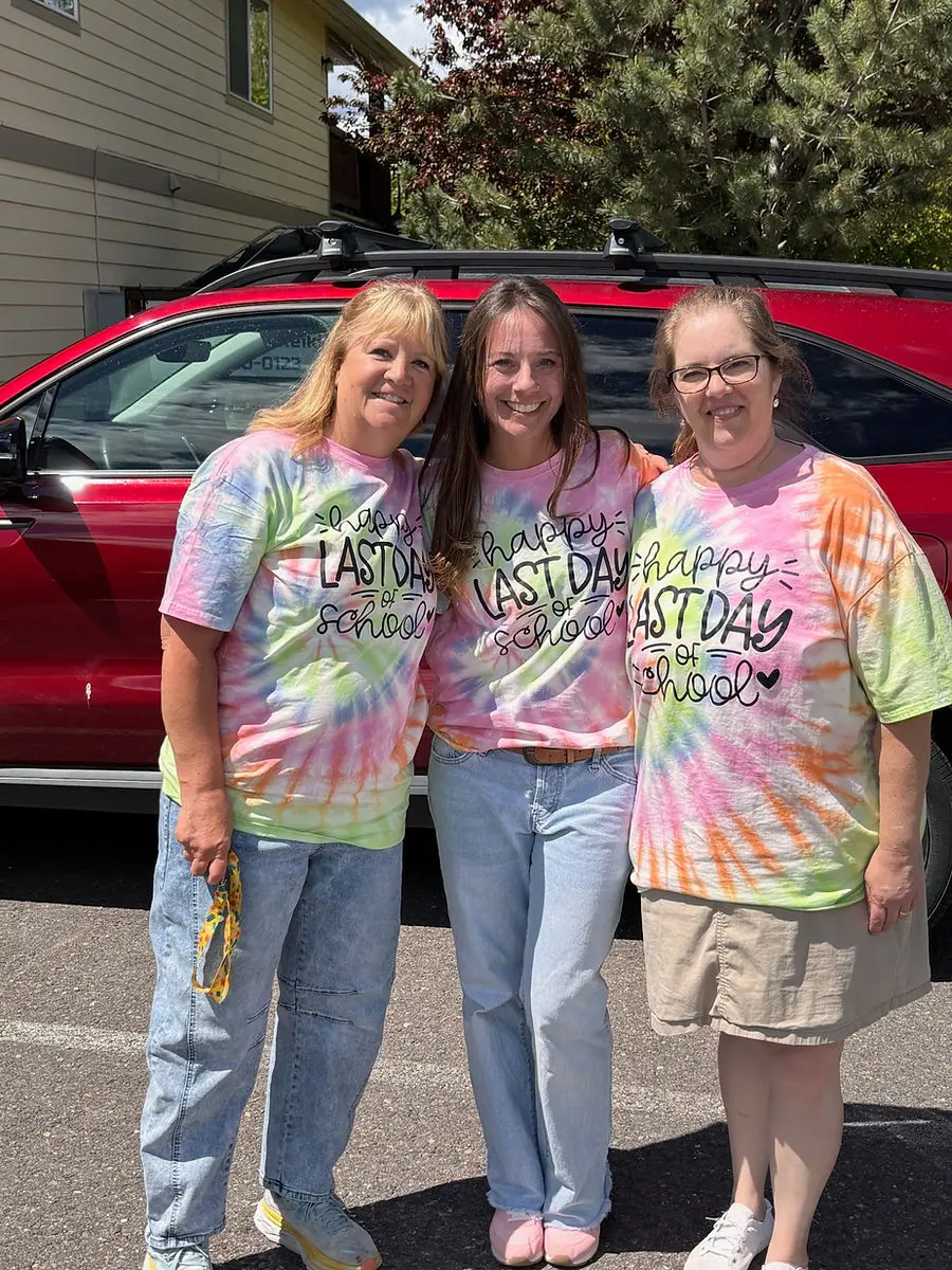 Three women stand together wearing matching tie-dye shirts that say "Happy Last Day of School."