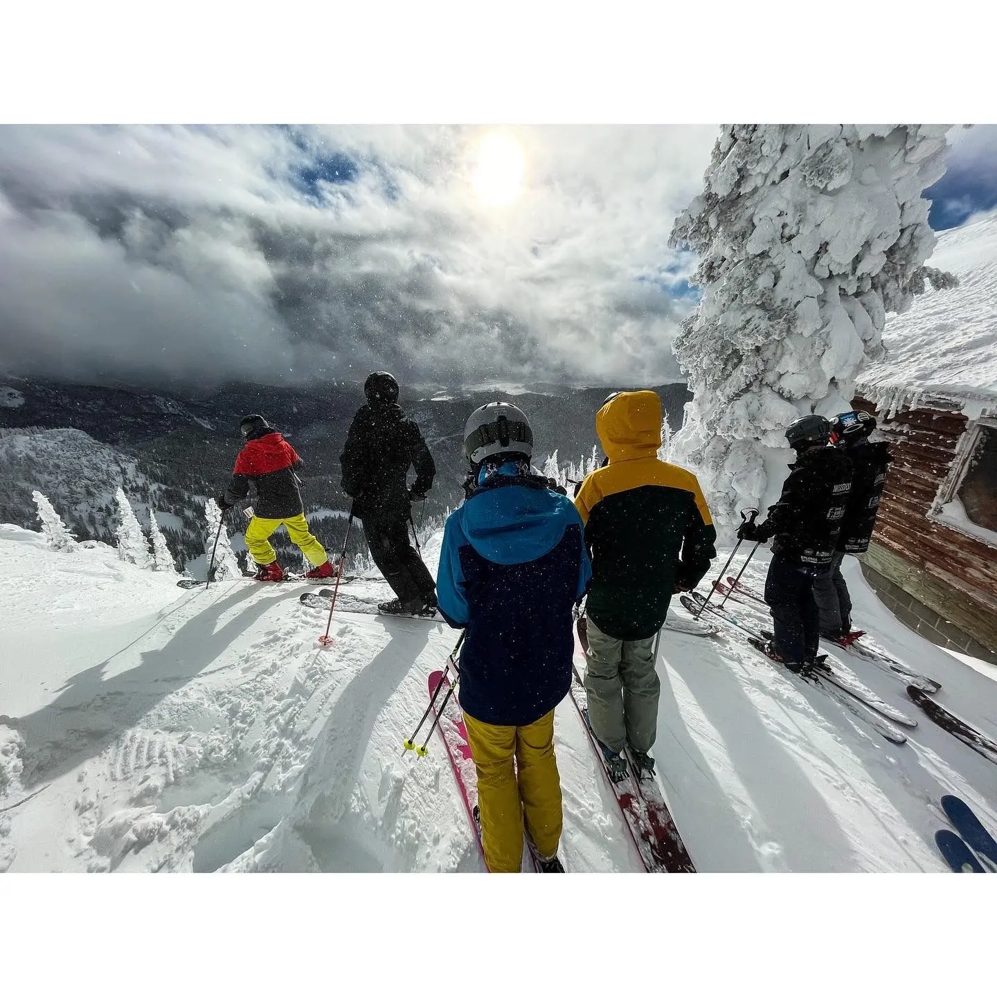 A group of people wearing ski gear stands on a snowy ridge overlooking a mountainous landscape.