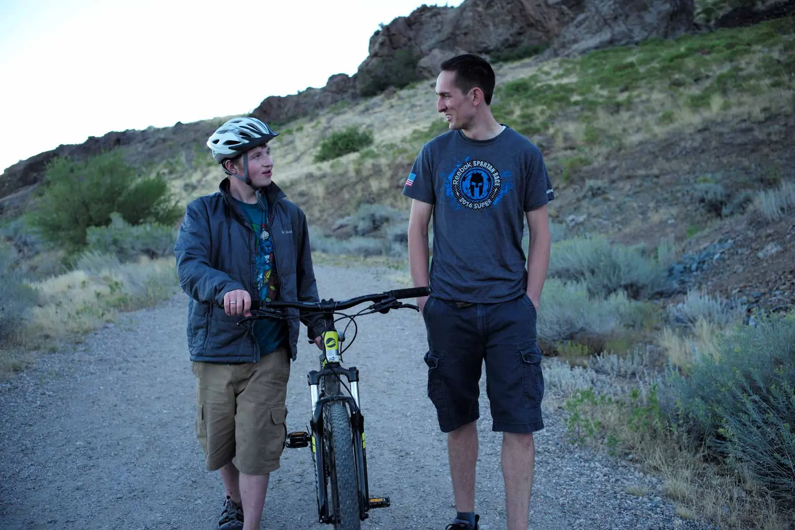 Two males walk together along a gravel path outdoors with a bicycle.