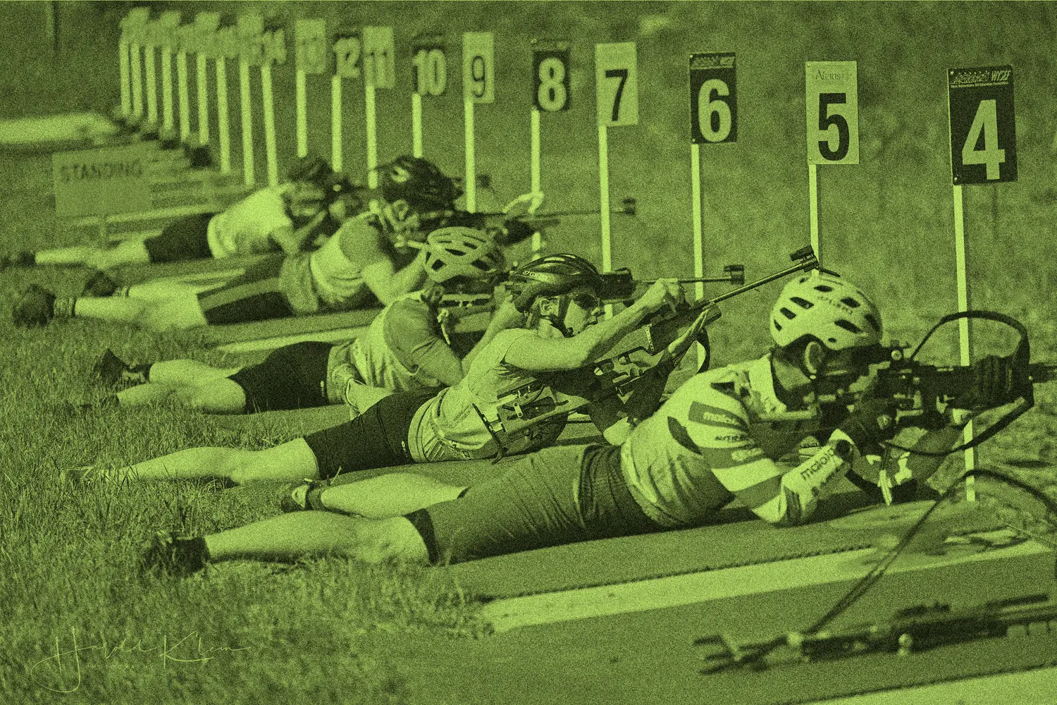 Athletes lie prone in a row while aiming rifles at numbered targets at a shooting range.