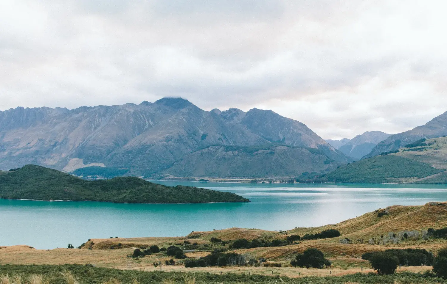 A scenic landscape shows a blue-green lake surrounded by hills and mountains under a cloudy sky.