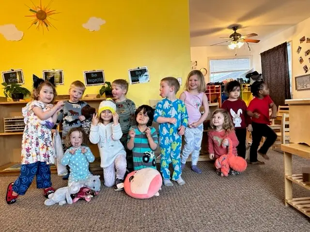 Group of young children in pajamas and casual clothes standing and sitting in a daycare or preschool classroom.