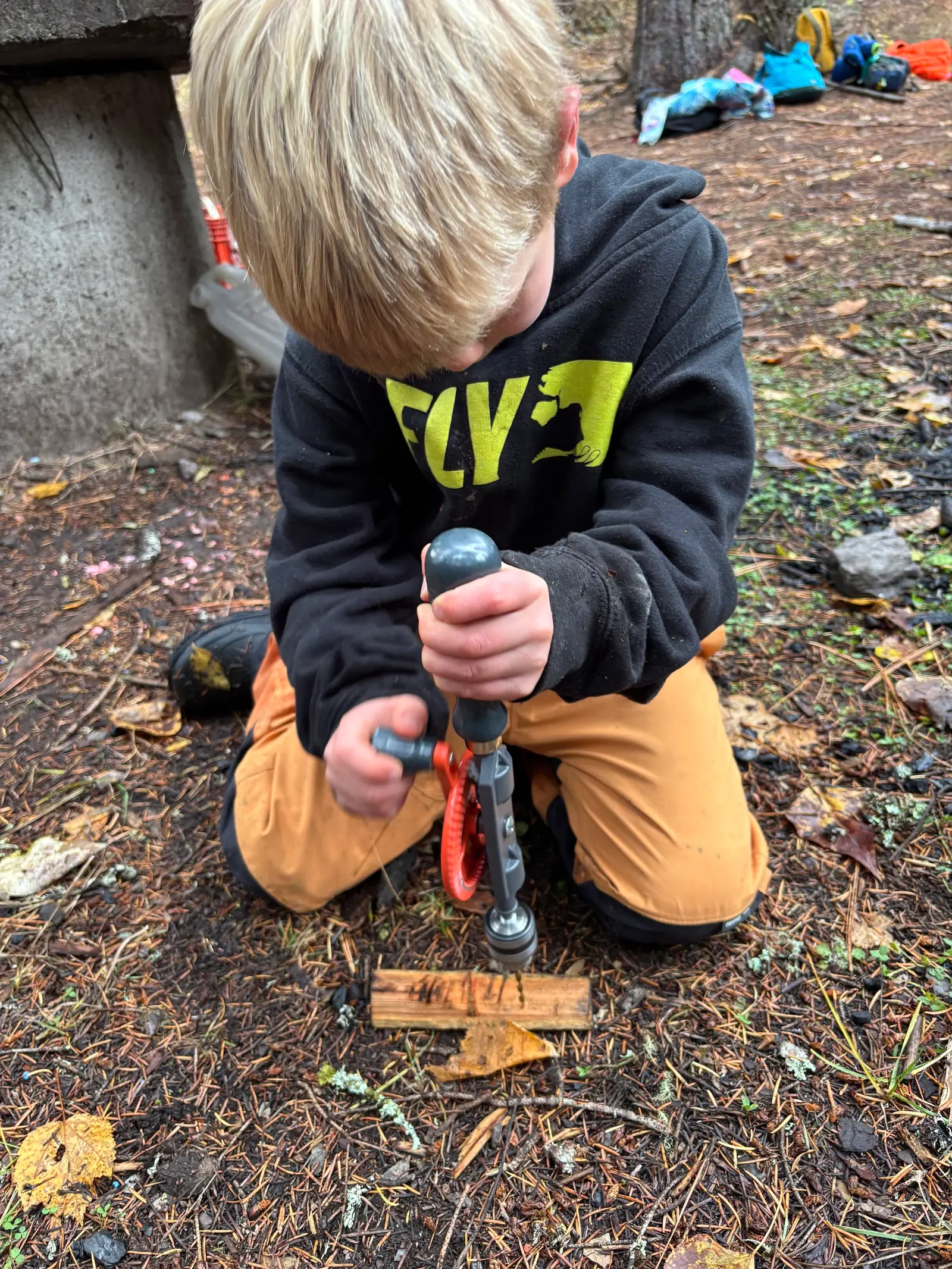 A child uses a manual hand drill on a piece of wood outdoors.