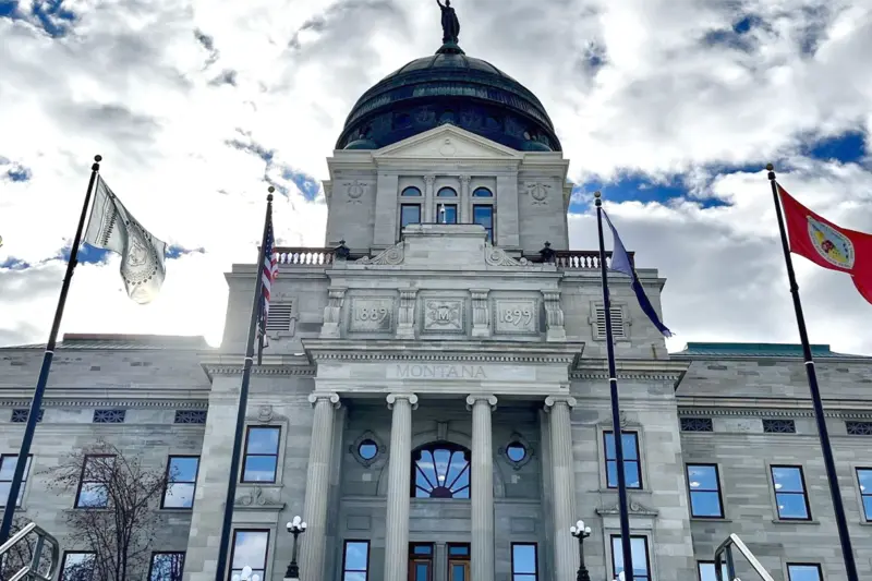 A photograph of the state capitol building with flags flying in front under a cloudy sky.