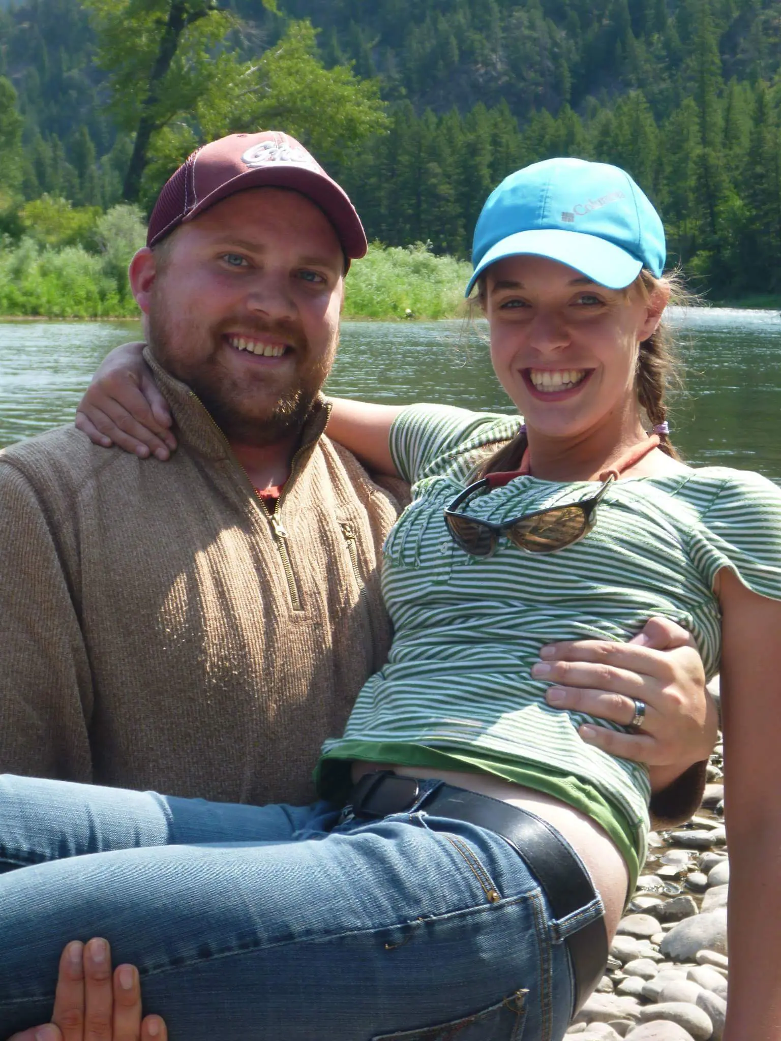 A man carries a woman in his arms while they stand near a river in front of a forest.
