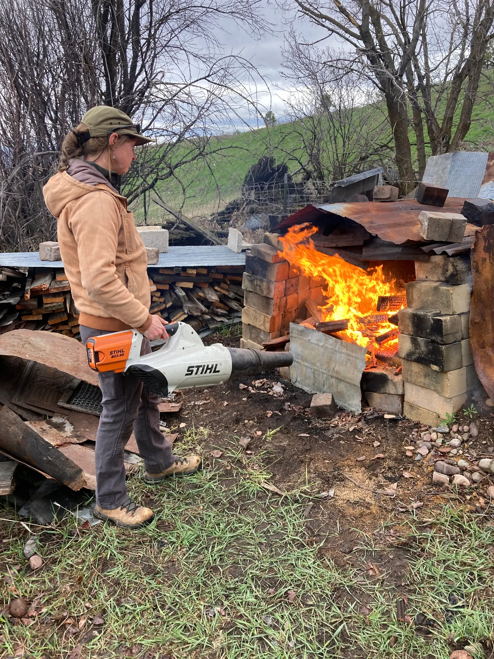 A person uses a leaf blower to stoke a fire in a cinder block fire pit.