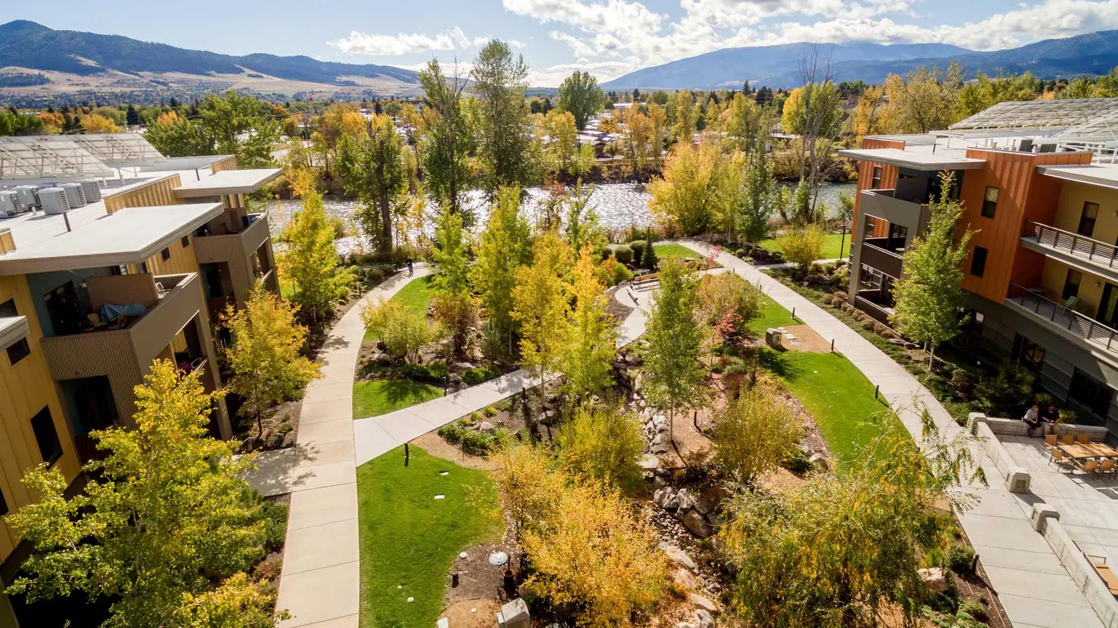 An elevated view of an apartment complex with a landscaped courtyard, river, and mountains in the background.