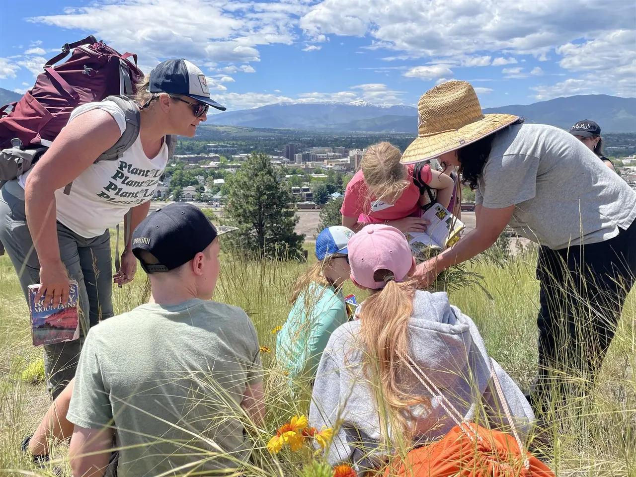An instructor shows a group of children something in a guidebook while they sit in a grassy field with a mountain view.