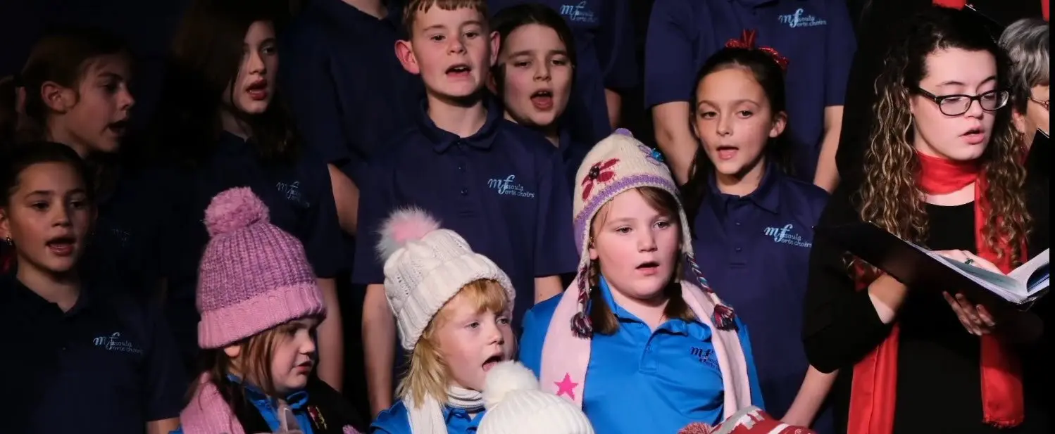 Children singing in a choir, some wearing winter hats and scarves, with a woman in the foreground holding a book and singing.