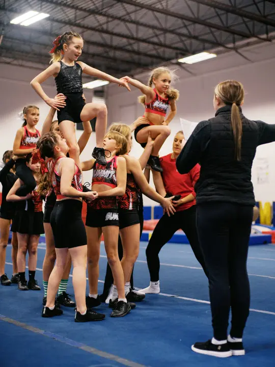 An instructor oversees a group of young cheerleaders practicing a stunt in a gym.