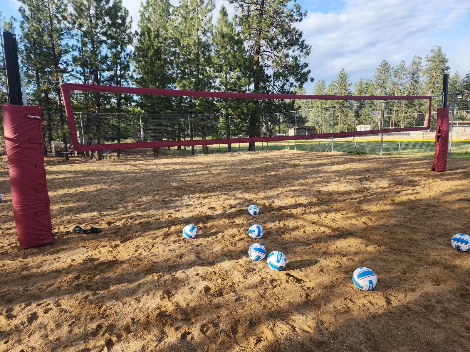 A volleyball net stands over a sandy court with several volleyballs scattered on the ground.