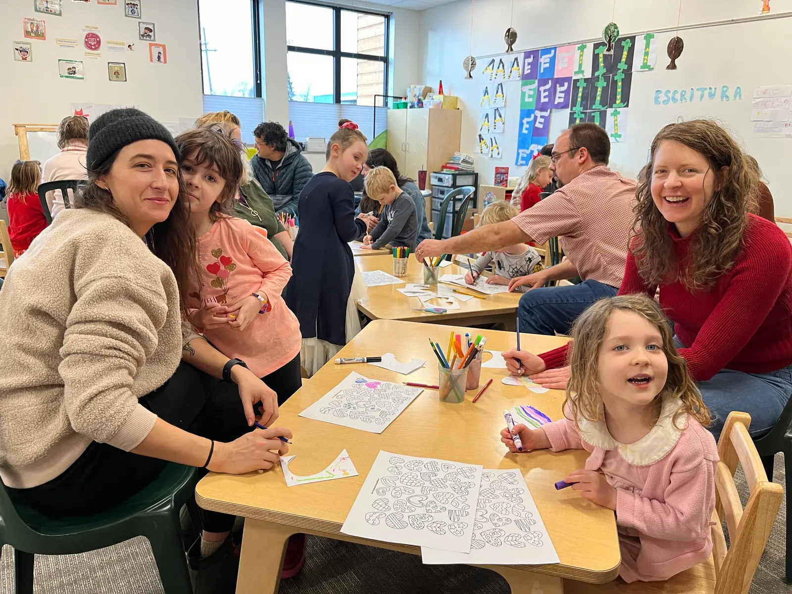Parents and children sit at tables together coloring worksheets in a classroom setting.
