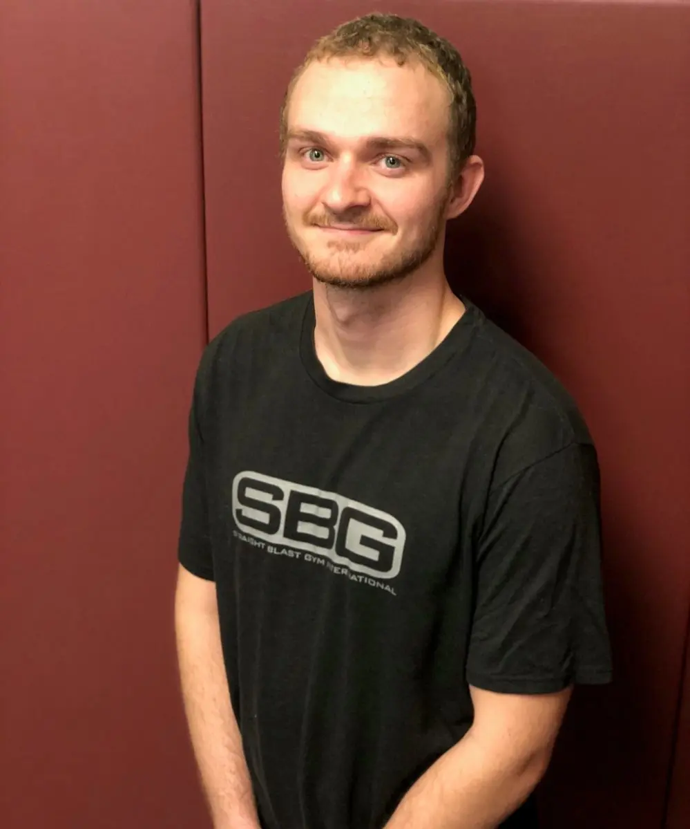 A man wearing a black shirt stands in front of a maroon wall.