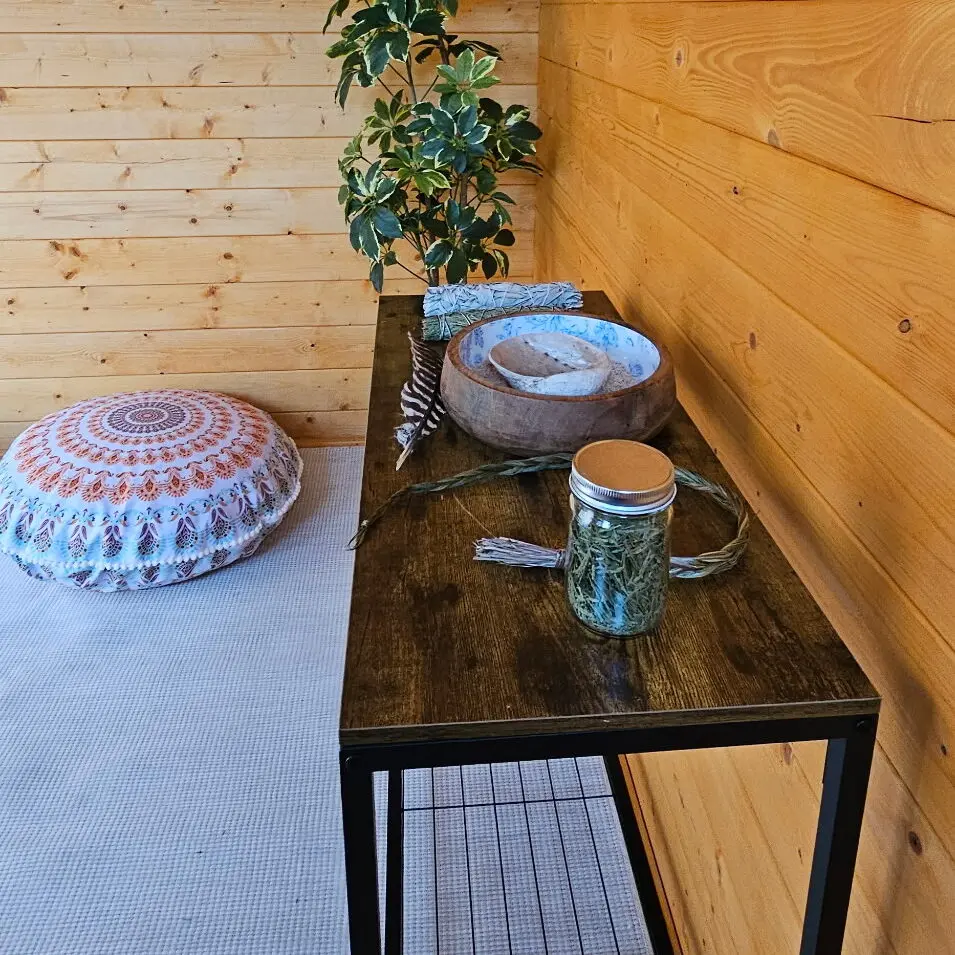 A wooden table with a jar of herbs, a bowl containing a shell, and a feather sits in front of a wood-paneled wall next to a patterned cushion.