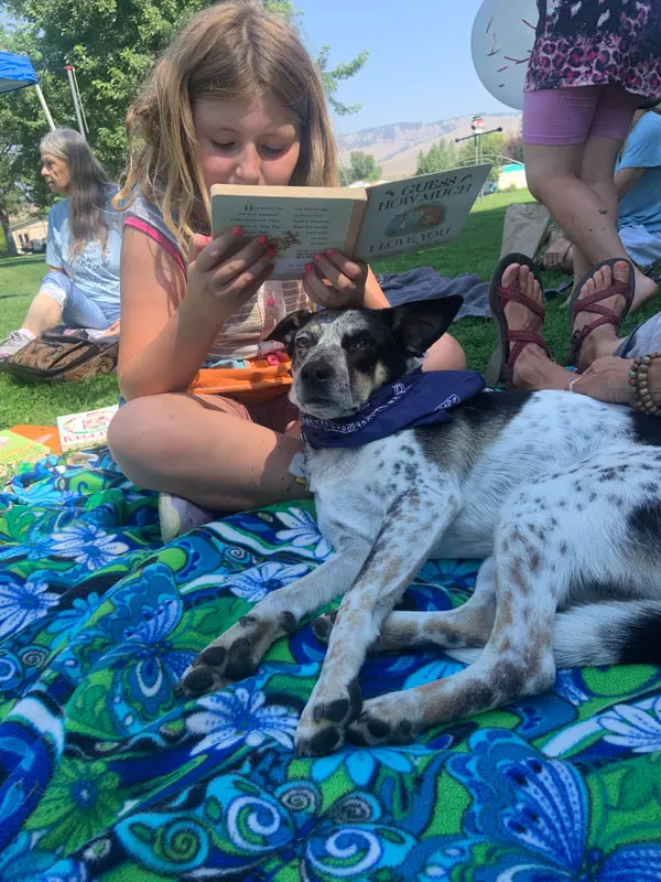 A young girl reads a book aloud while sitting on a blanket next to a dog wearing a blue bandana.