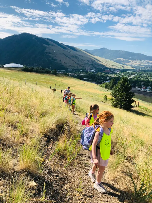 A group of children and an adult hike along a hillside trail with mountains in the background.