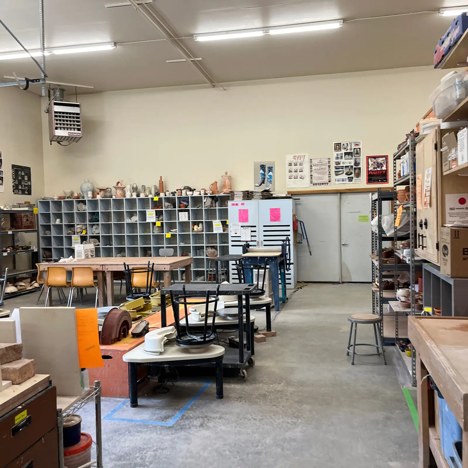 The image shows a pottery studio filled with work tables, clay storage cubbies, and pottery wheels.