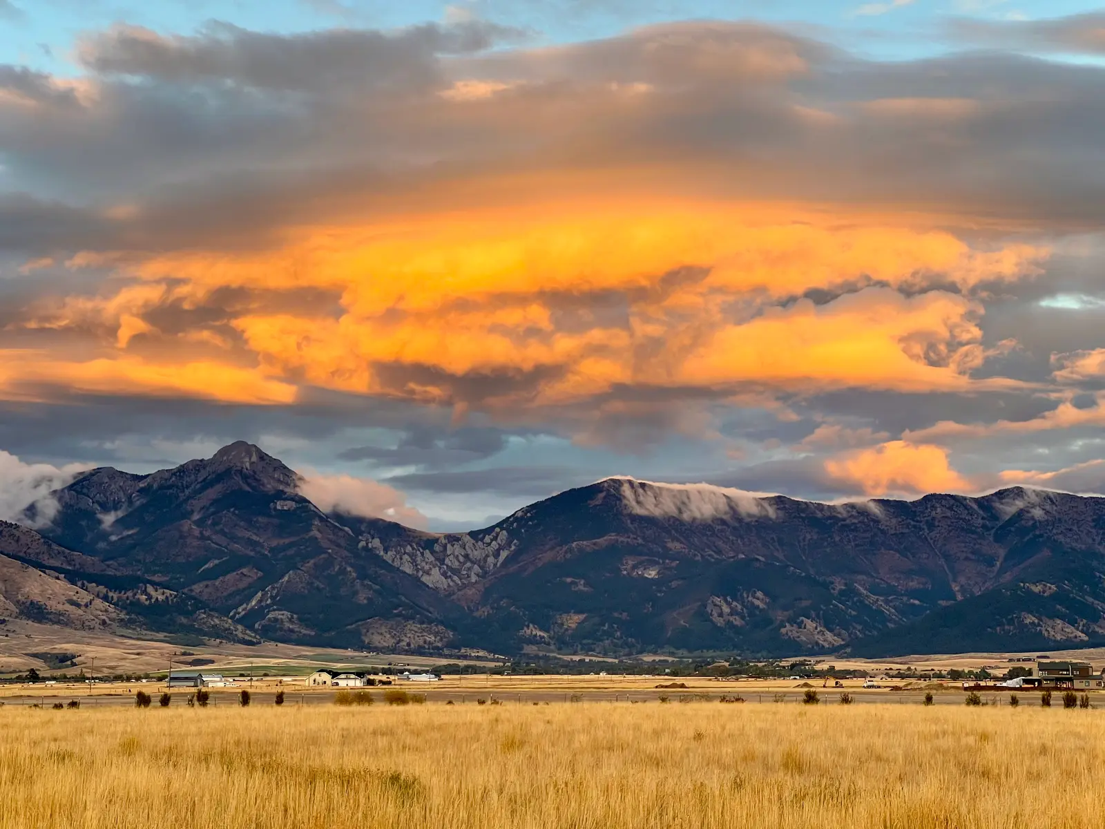 A scenic view of a mountain range under a glowing orange sunset sky with a golden grass field in the foreground.