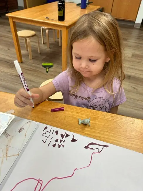 A young girl sits at a table and draws with a purple marker.