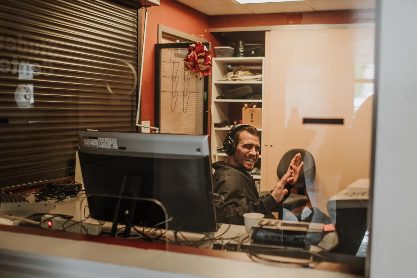 A man wearing headphones is smiling and gesturing inside a radio or recording studio.