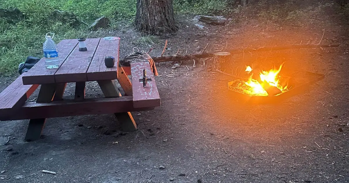 A campfire burns next to a wooden picnic table at a campsite.