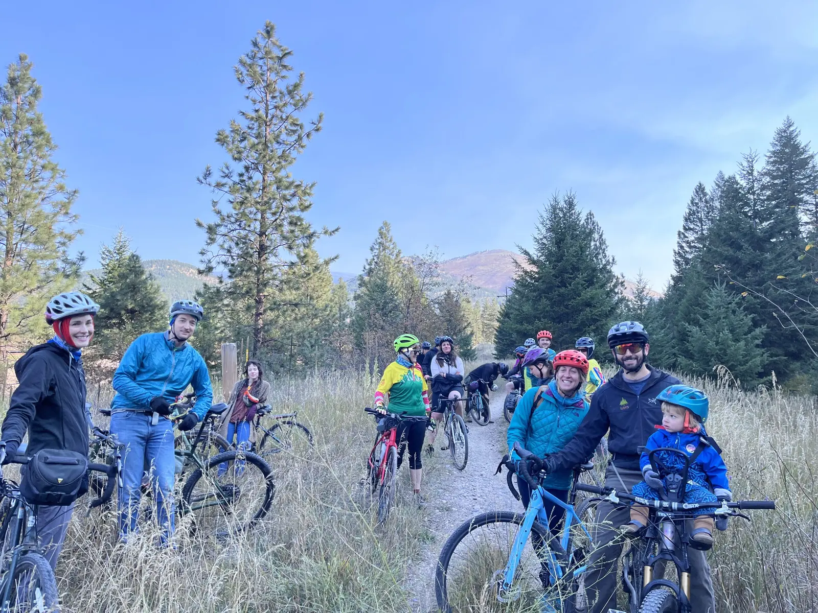 A group of cyclists including adults and a small child in a seat on a bike take a break on a dirt trail.