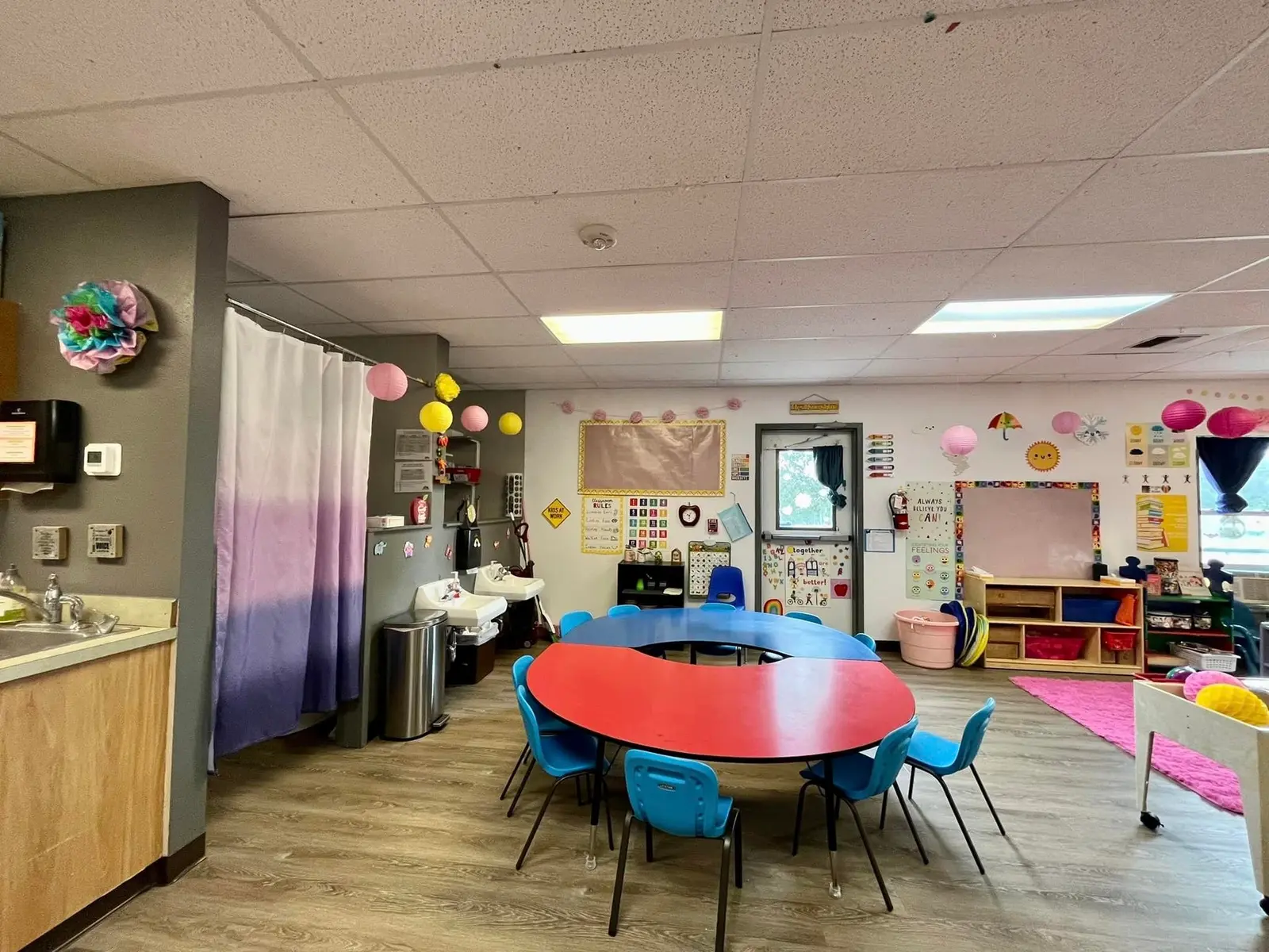 A view of an indoor preschool classroom featuring a central red and blue circular table surrounded by blue chairs.
