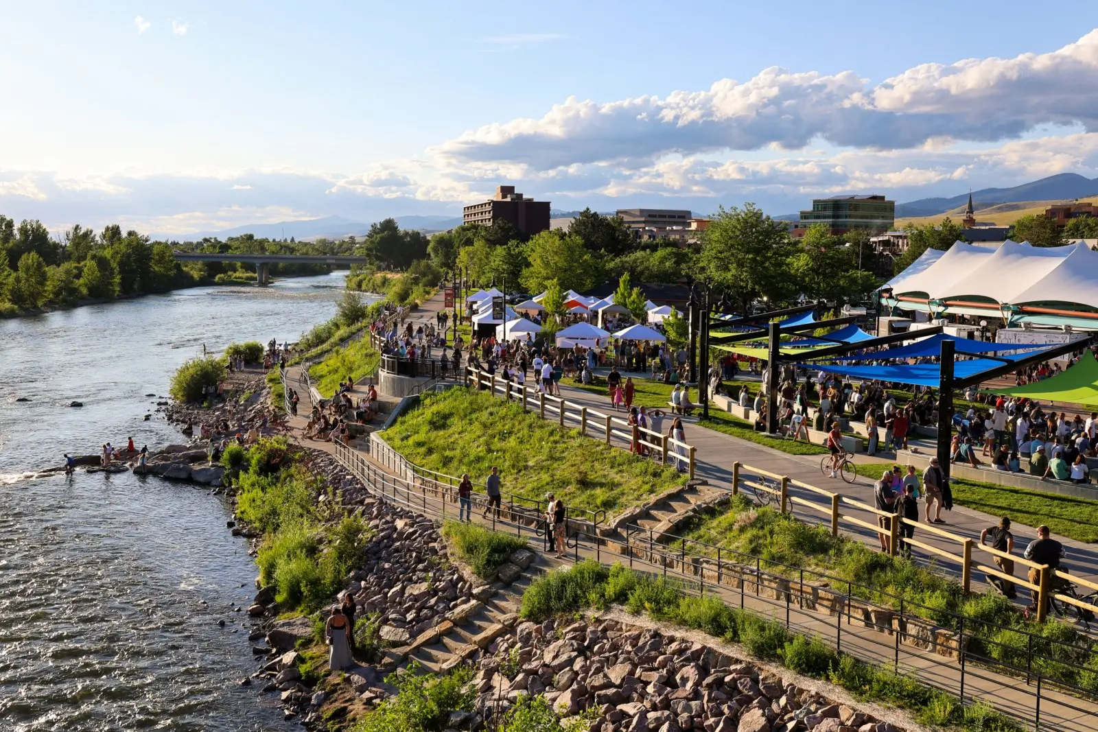 A riverfront path is bustling with people visiting white event tents and a large canopy structure at sunset.