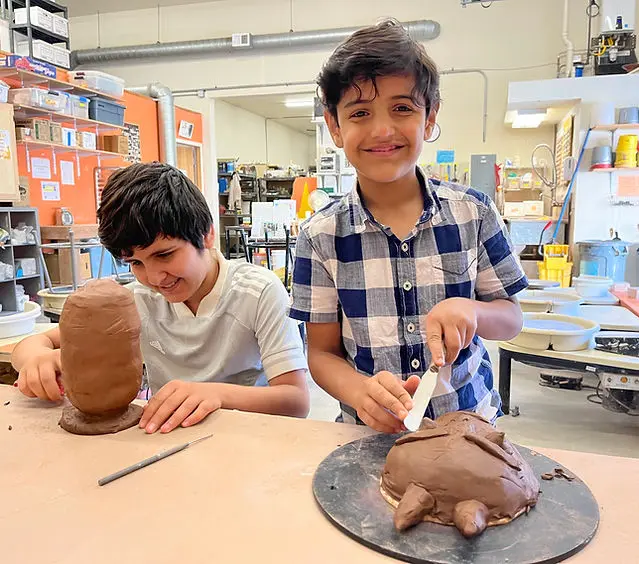 Two young boys working on clay projects. One with dark hair, darker skin, and a gray tshirt works on the body of an in-progress piggy bank, while the other boy who looks related to the first and wears a blue gingham shirt holds up a tool to a mask he is making while smiling for the camera.