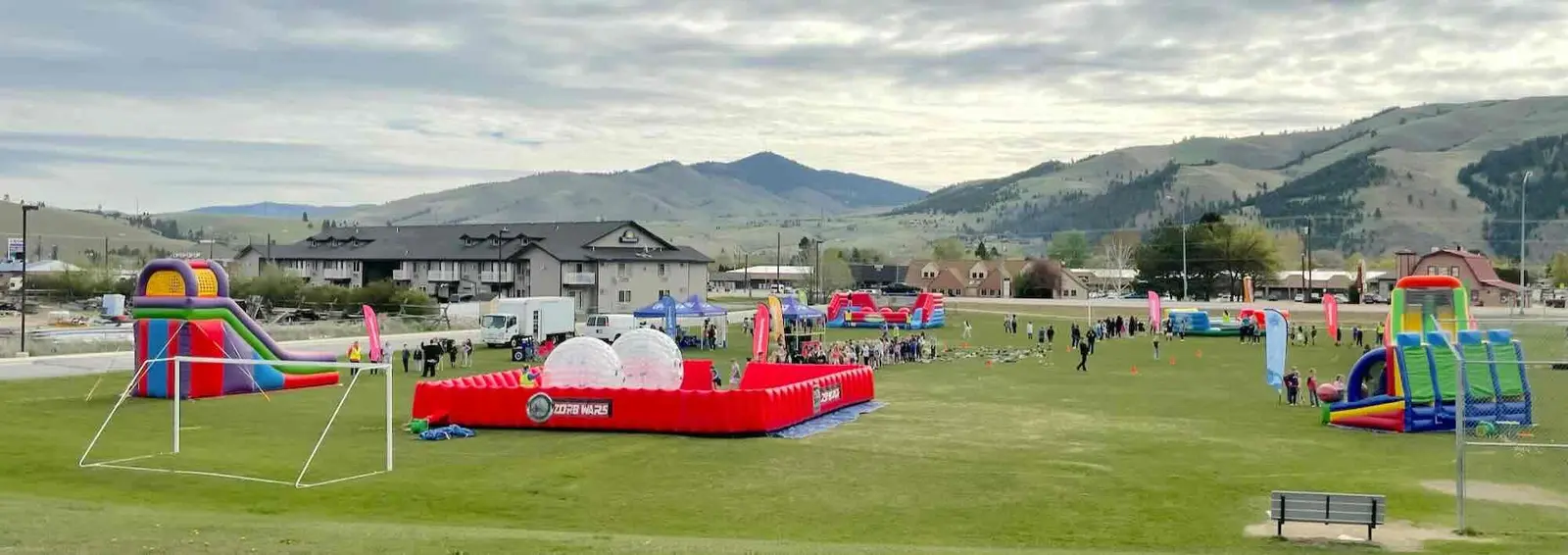 People are gathered on a grassy field filled with inflatable activities including a Zorb Wars arena and bounce houses.