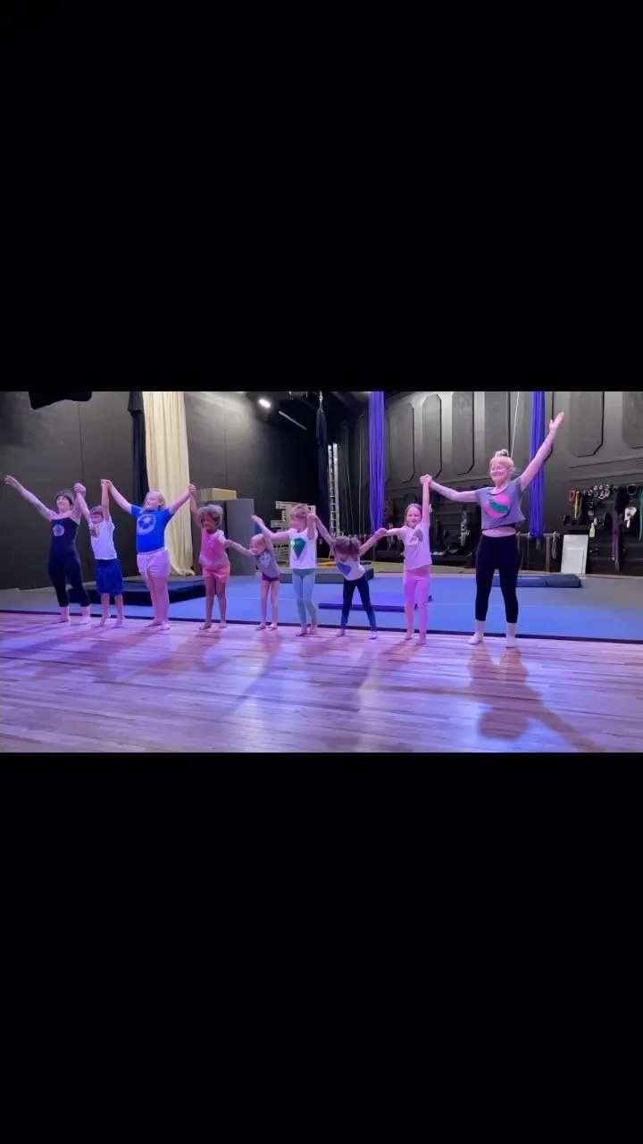 A group of children and an instructor stand in a line holding hands in an exercise studio.