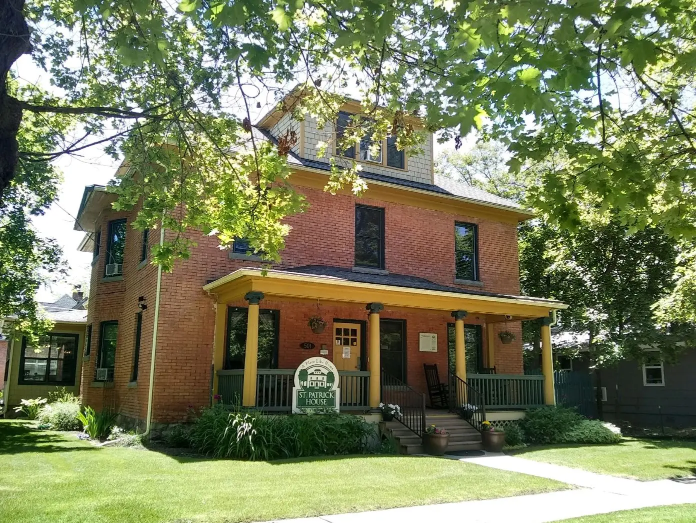 A brick house with a front porch is surrounded by trees and a green lawn.