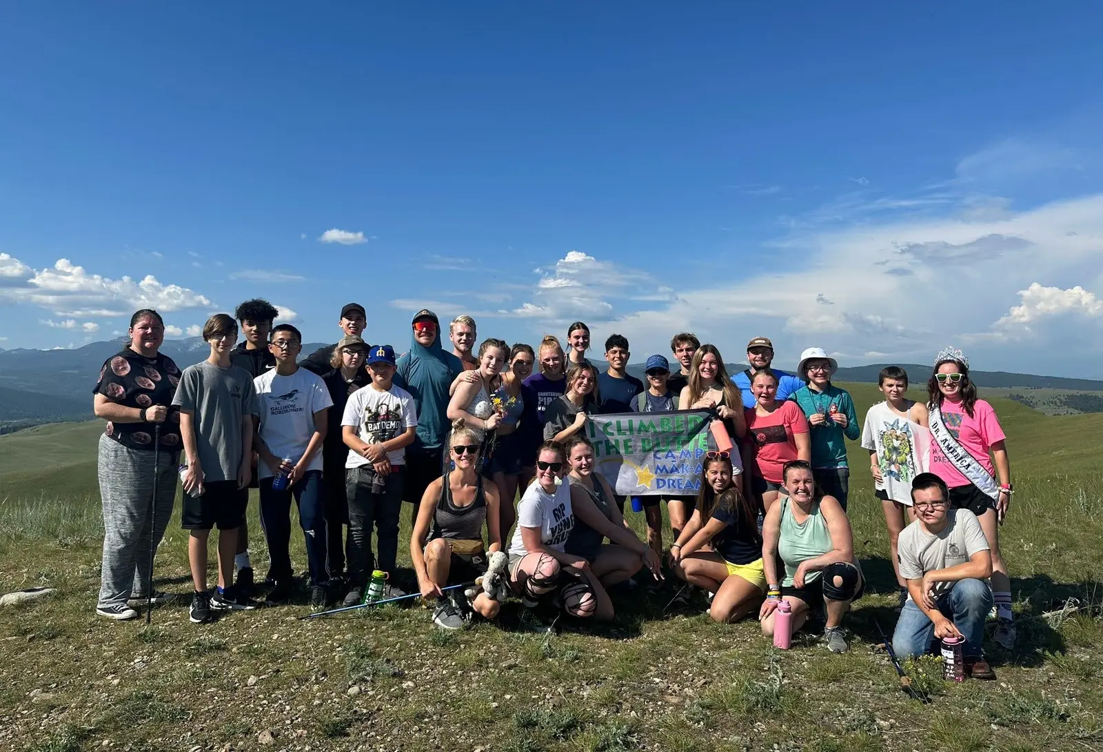 A large group of young people stands together on a grassy hillside after hiking.