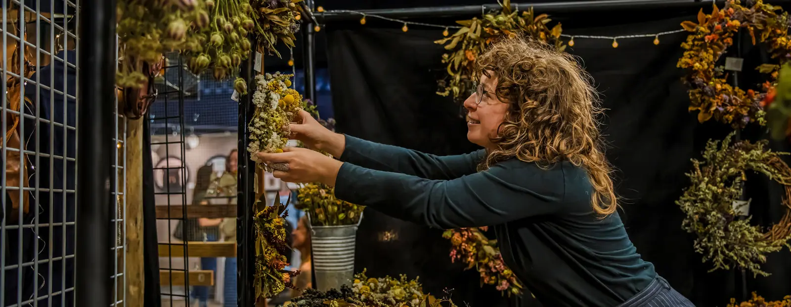 A woman with curly hair adjusts a dried flower arrangement at a market stall.