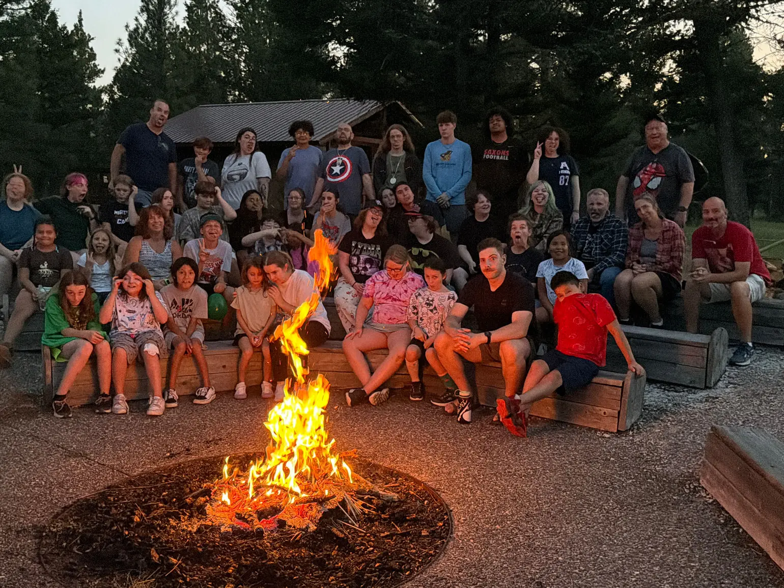 A large group of people are gathered around a campfire at dusk.
