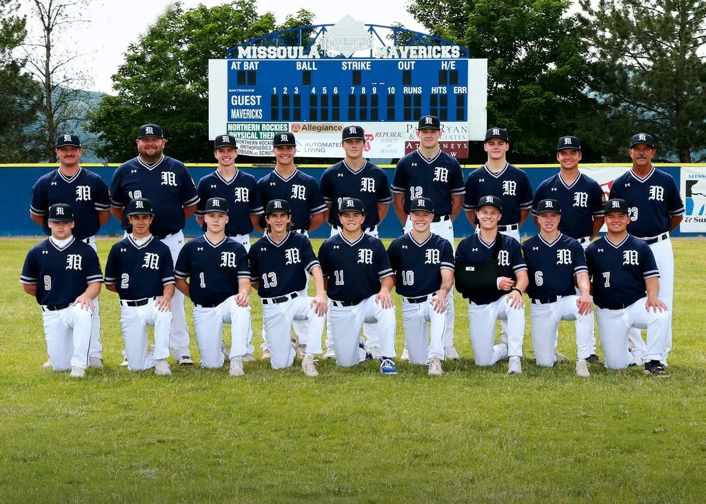 A baseball team poses for a group photograph on a field in front of a scoreboard.