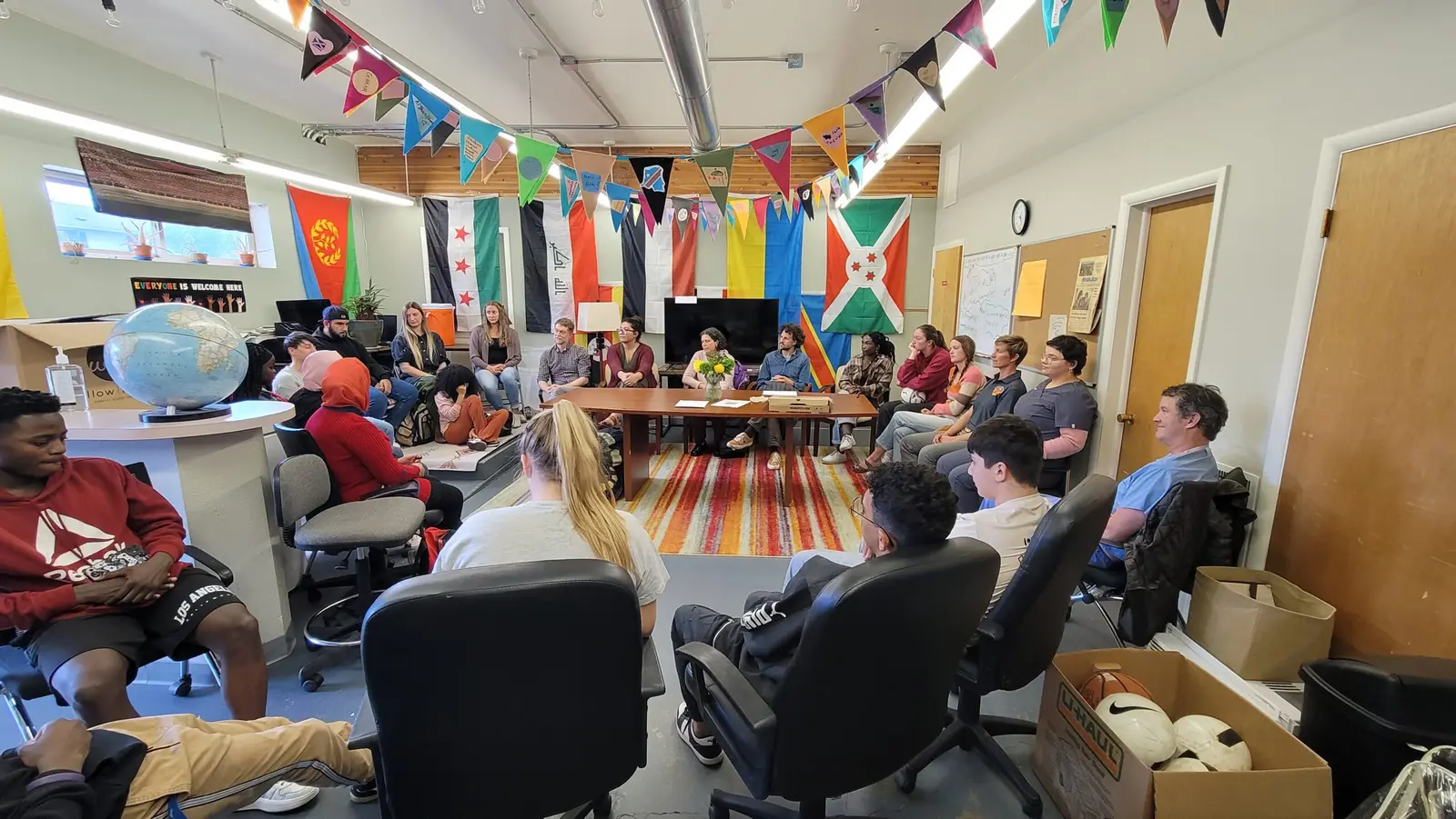 A diverse group of people gathers in a meeting room decorated with various flags.