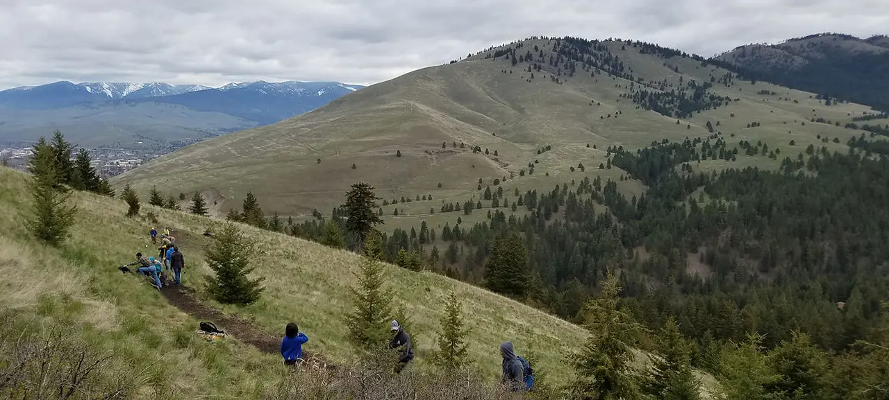 A group of people hikes along a trail on a grassy hillside with mountain ranges in the distance.