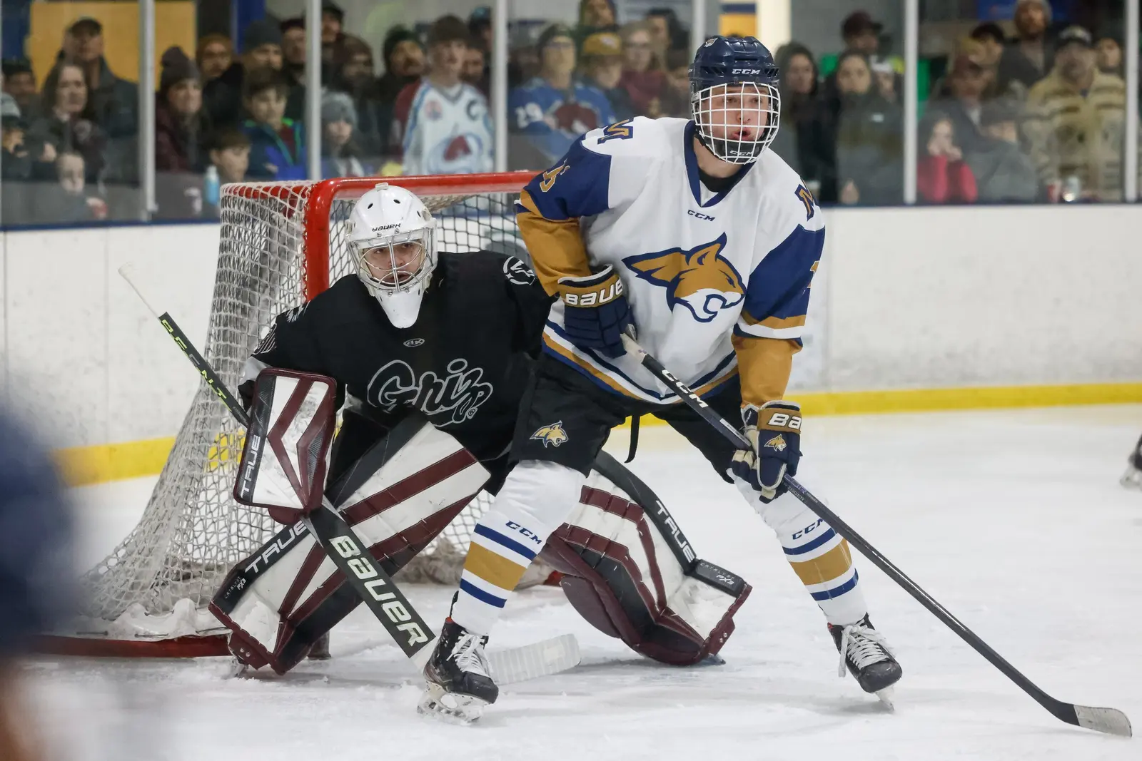 An ice hockey player in a white and blue jersey positions himself in front of a goaltender at a game.