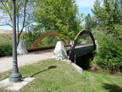 Photo from oza.com A wooden arched bridge crosses over a small stream surrounded by green trees and foliage.
