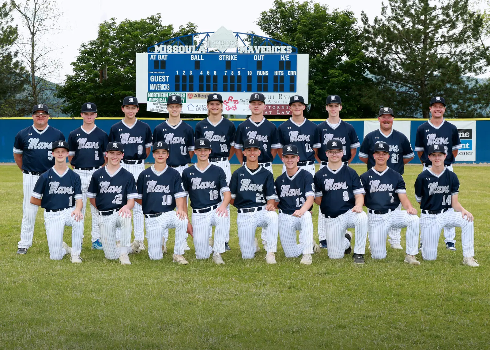 A baseball team poses for a group photograph on a field in front of a scoreboard.