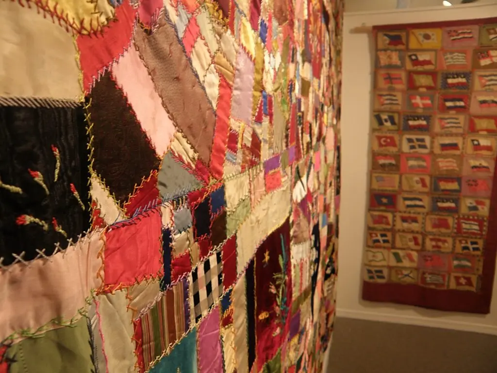 A close-up view of a large patchwork quilt displayed alongside a flag-patterned wall hanging in a gallery.