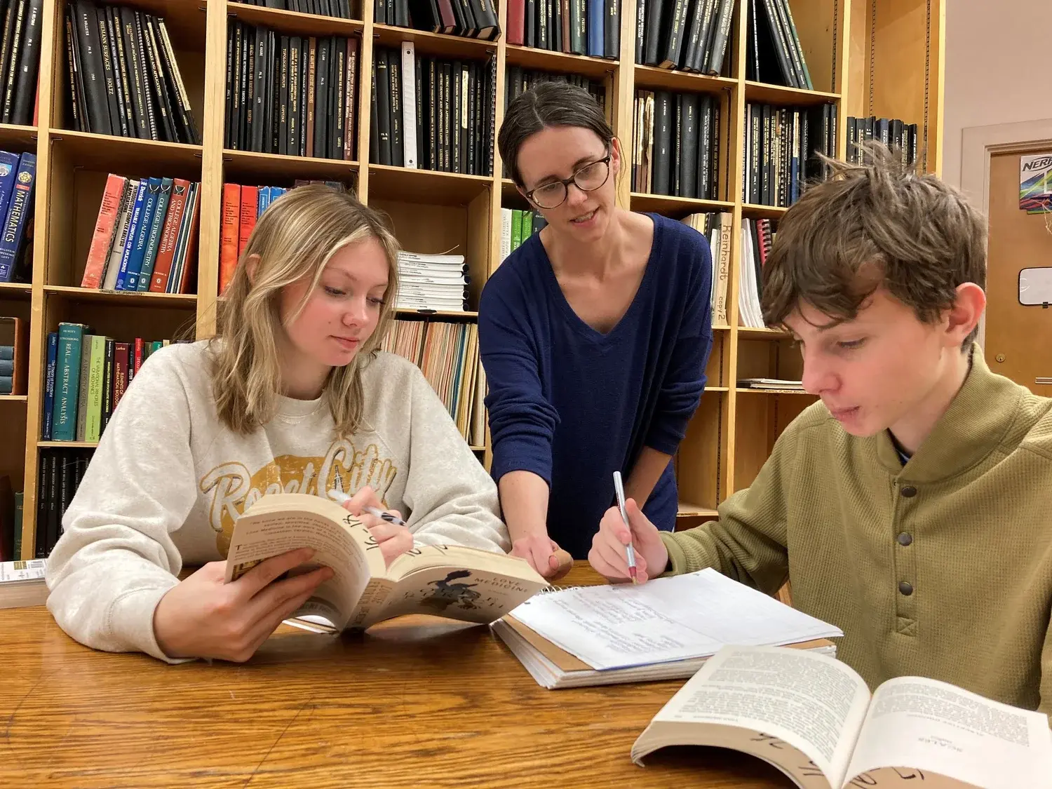 A teacher helps two students study at a table in front of a large bookshelf.