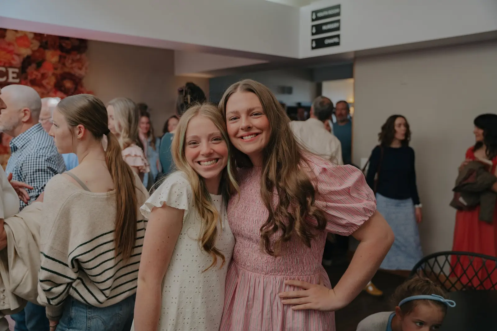 Two young women smile for the camera in a room filled with other people.