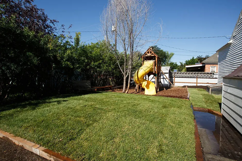 A backyard features a wooden play set with a yellow slide on a lawn.